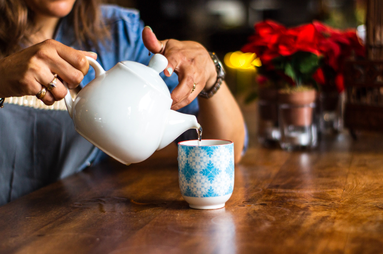 A woman pours tea from a white ceramic teapot into a small ceramic glass sitting on a table.
