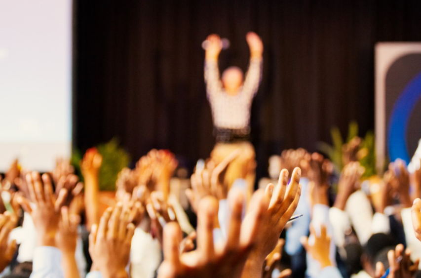 A large crowd raises their hands in front of a speaker on stage.