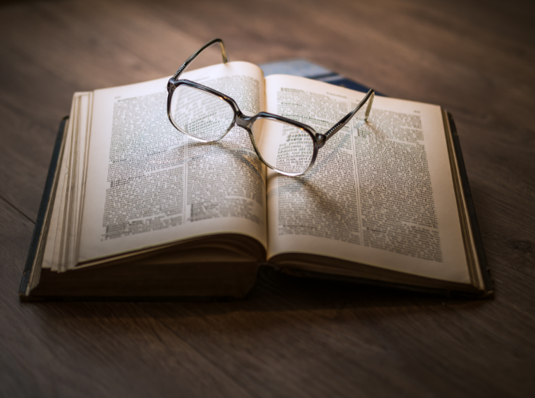 A brown-rimmed pair of glasses sit on top of a thick, open book.