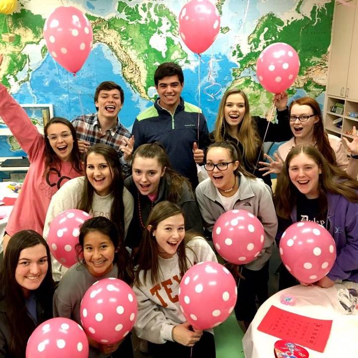 A group of smiling students kneel in a classroom holding pink and white spotted balloons.
